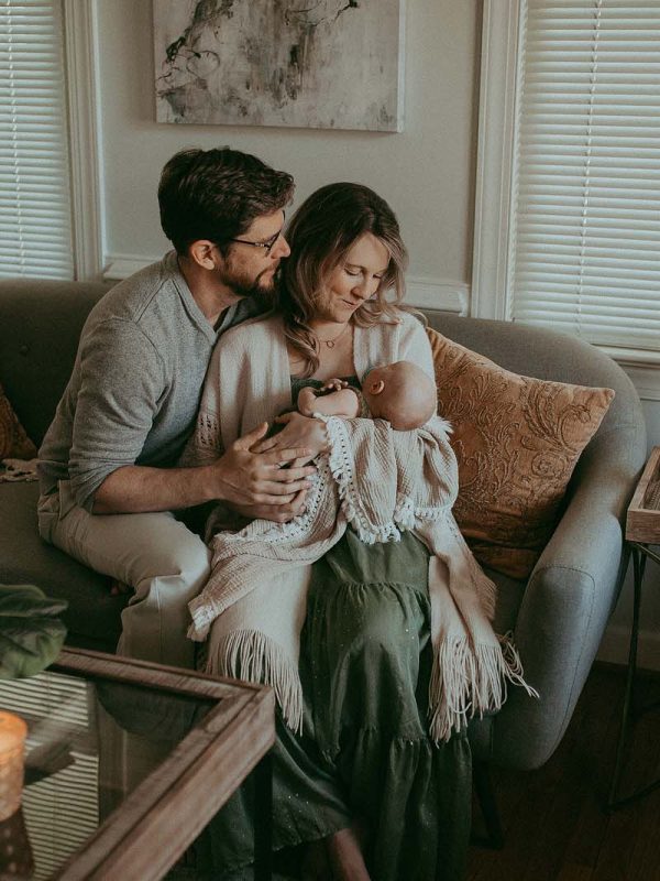A sweet moment captured during an in-home newborn photo session, showing the new mom wearing a green maxi dress and holding her baby boy in her arms while the dad sits next to them, looking on with a loving smile. The portrait was taken by Cary Newborn Photographer - Victoria Vasilyeva Photography