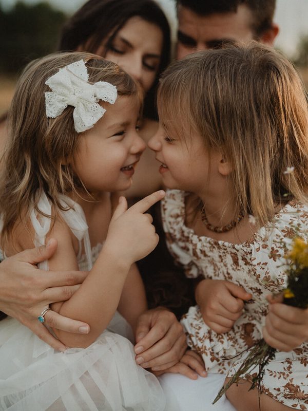 Close-up of two sisters sharing a sweet moment outdoors, touching noses while sitting on a blanket. The younger girl points playfully at her sister's face while holding yellow wildflowers, with their parents watching affectionately in the soft-focus background. Family photo session in Raleigh with Victoria Vasilyeva Photography.