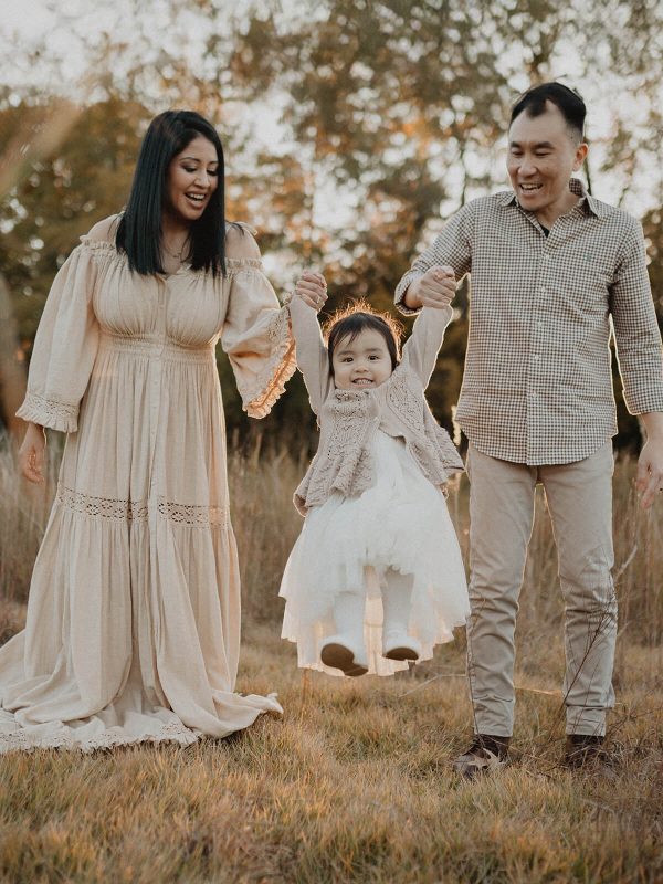 Smiling parents holding their toddler daughter by the hands and lifting her in the air during an outdoor family photo session in a golden field at Anderson Point Park in Raleigh. Photo by Victoria Vasilyeva Photography, family photographer in Raleigh.