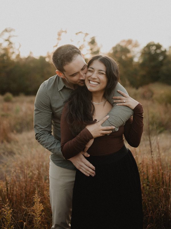 Beautiful couple embracing and laughing during a family photo session at sunset in a grassy field in Raleigh, North Carolina. Natural light family photography by Victoria Vasilyeva Photography.