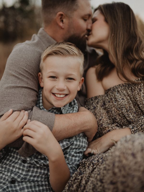 Young boy smiling at the camera while his parents share a kiss behind him during an outdoor family photo session in Raleigh, NC. Family photography by Victoria Vasilyeva Photography.