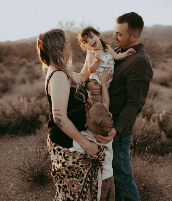 Family standing on a field at sunset, dad holding toddler girl while her brother standing next to them and putting hands up to her. Portrait by Victoria Vasilyeva Photography – family photographer in Raleigh, North Carolina.