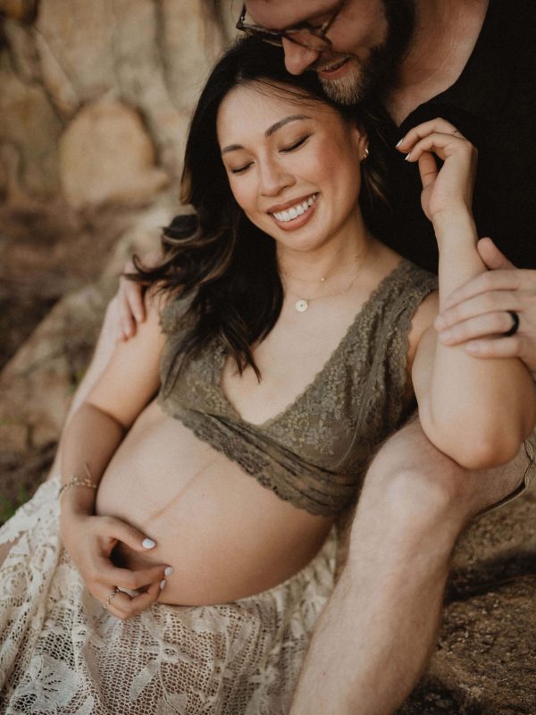 Close and romantic moment of a couple holding hands and embracing near rocks, focusing on the expecting mother and her baby bump. Maternity photography in Raleigh, NC by Victoria Vasilyeva Photography.