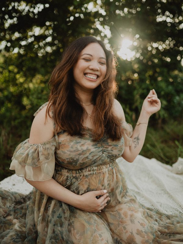 Pregnant woman smiling and holding her baby bump during an outdoor maternity photo session with soft sunlight near Raleigh, North Carolina. Natural maternity photography by Victoria Vasilyeva Photography.