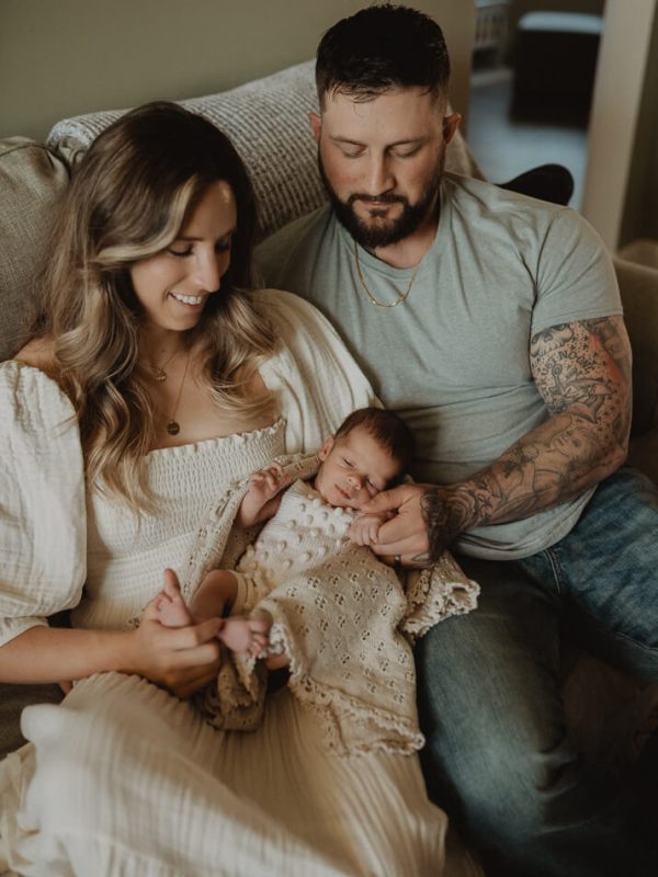 Family sitting together on the couch during an in-home newborn session, with mom holding the baby boy wrapped in a textured blanket. Lifestyle newborn photography by Victoria Vasilyeva Photography – Cary, NC newborn photographer.