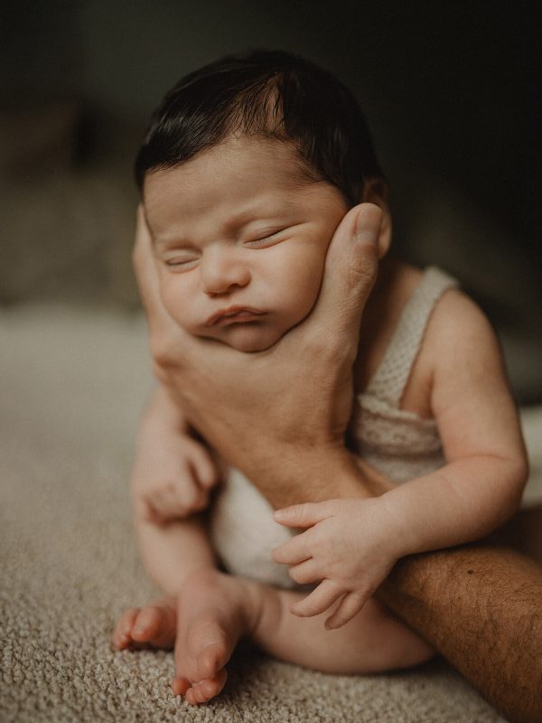 Newborn baby sitting upright while sleeping and being supported by a dad’s hand, dressed in a beige knit outfit. Portrait by Victoria Vasilyeva Photography – newborn photographer in Cary, NC.