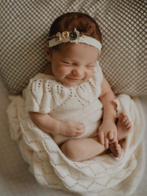 Smiling newborn baby girl wearing a floral headband and cream knitted outfit, posed on a soft blanket with her feet curled up. Photographed by Victoria Vasilyeva Photography, newborn photographer in Cary, NC.