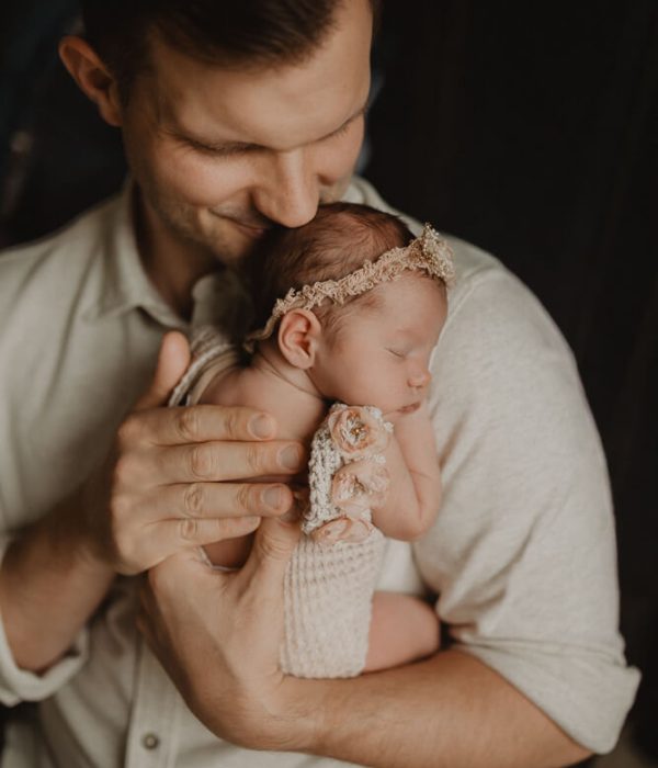 Father holding newborn baby girl close to his chest, cuddling her while she sleeps in a knitted wrap and headband. Photographed by Victoria Vasilyeva Photography, newborn photographer in Cary, NC.