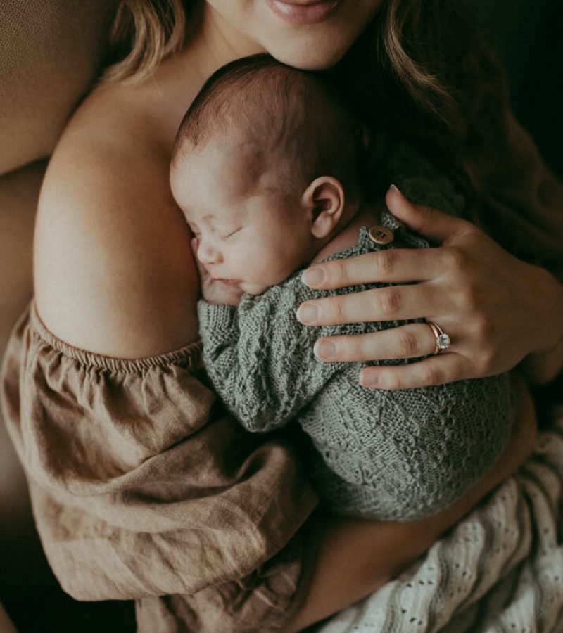 The portrait of newborn boy sleeping on mom's chest. He wears knitted green romper and has dark short hair. The photo was taken by Raleigh newborn photographer - Victoria Vasilyeva Photography.