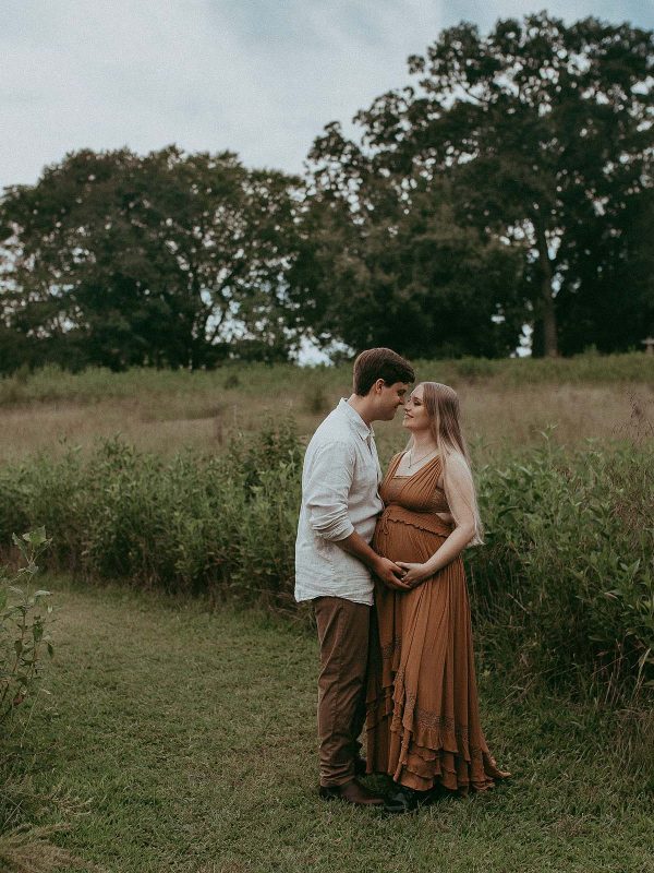 In a picturesque outdoor setting, an expecting mother with flowing blonde locks elegantly showcases her pregnancy glow, dressed in a boho maxi dress, while being photographed by a talented Raleigh maternity photographer in a field of tall grass.
