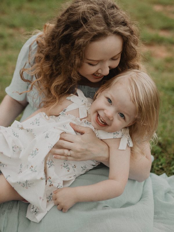 Mother with blonde curly hair holding her daughter in her lap while they laugh together outdoors. Photo by Victoria Vasilyeva Photography – Raleigh NC lifestyle family photographer.