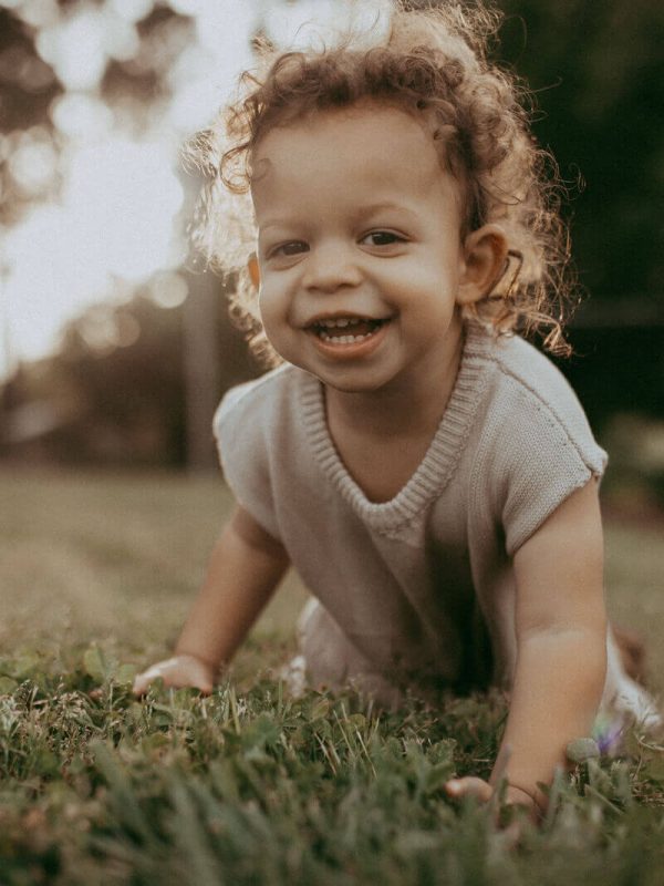 Close-up of a happy toddler boy with curly hair crawling on the grass at sunset. Captured by Victoria Vasilyeva Photography – Raleigh NC family photographer.