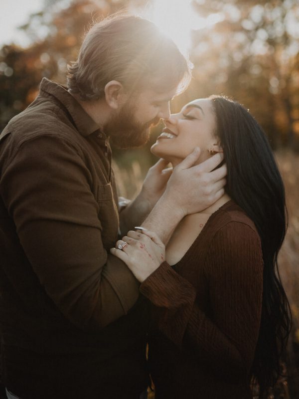Mom with black long hair and dad embracing and smiling at sunset while holding each other in a field with warm golden light. Image captured by Victoria Vasilyeva Photography – family and couples photographer in Raleigh, North Carolina.