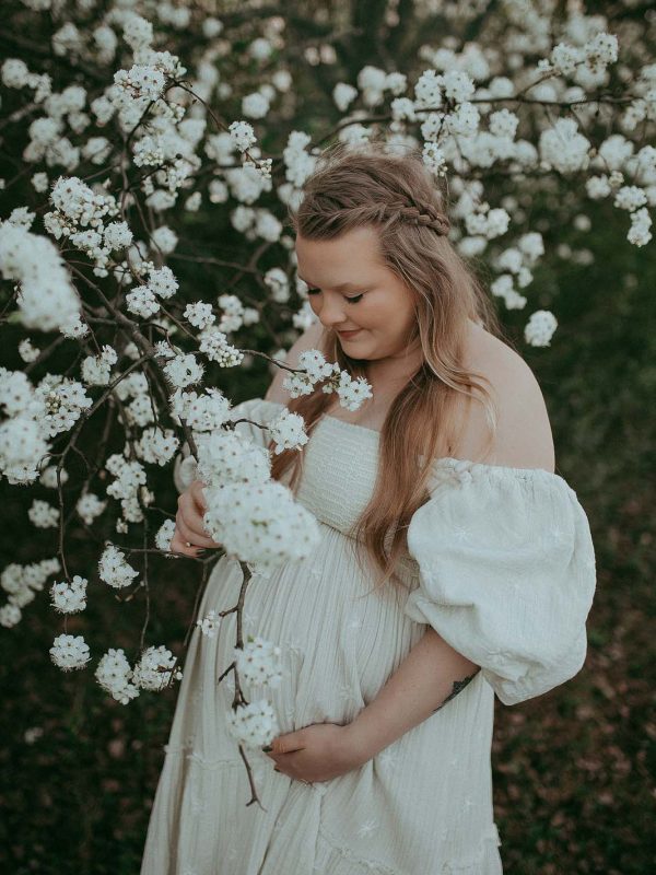In a picturesque setting among blooming trees, a mom-to-be with long blonde hair showcases her boho style in a flowing dress, captured by a talented Raleigh maternity photographer.