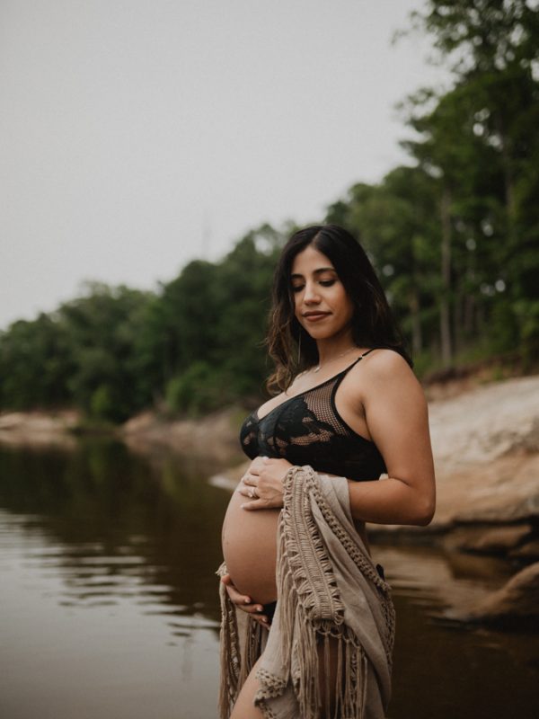 Pregnant woman with black long hair wearing a black bra and neutral-toned cover up and holding her belly during an outdoor maternity session in Raleigh, NC. Maternity photography by Victoria Vasilyeva Photography.