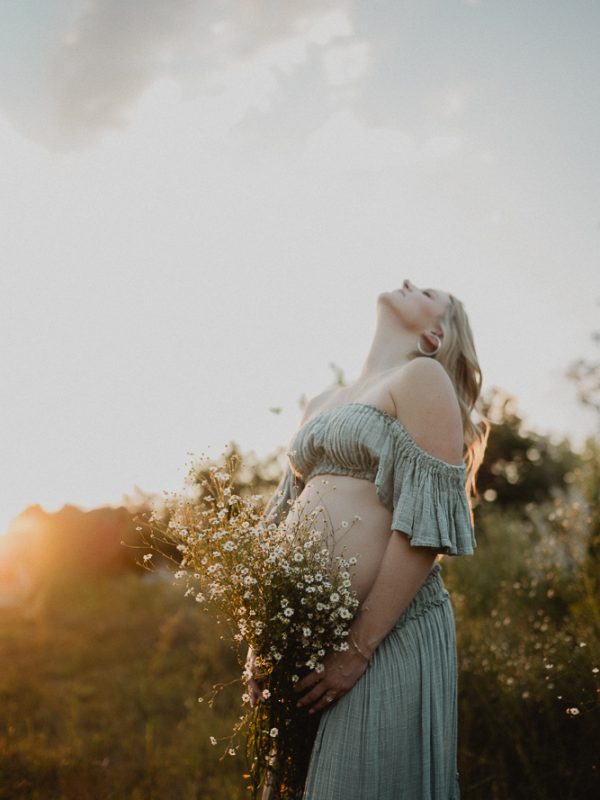 Pregnant woman holding a bouquet of white and yellow wildflowers at sunset. She is standing in an open field. Maternity photography in Raleigh NC by Victoria Vasilyeva Photography.